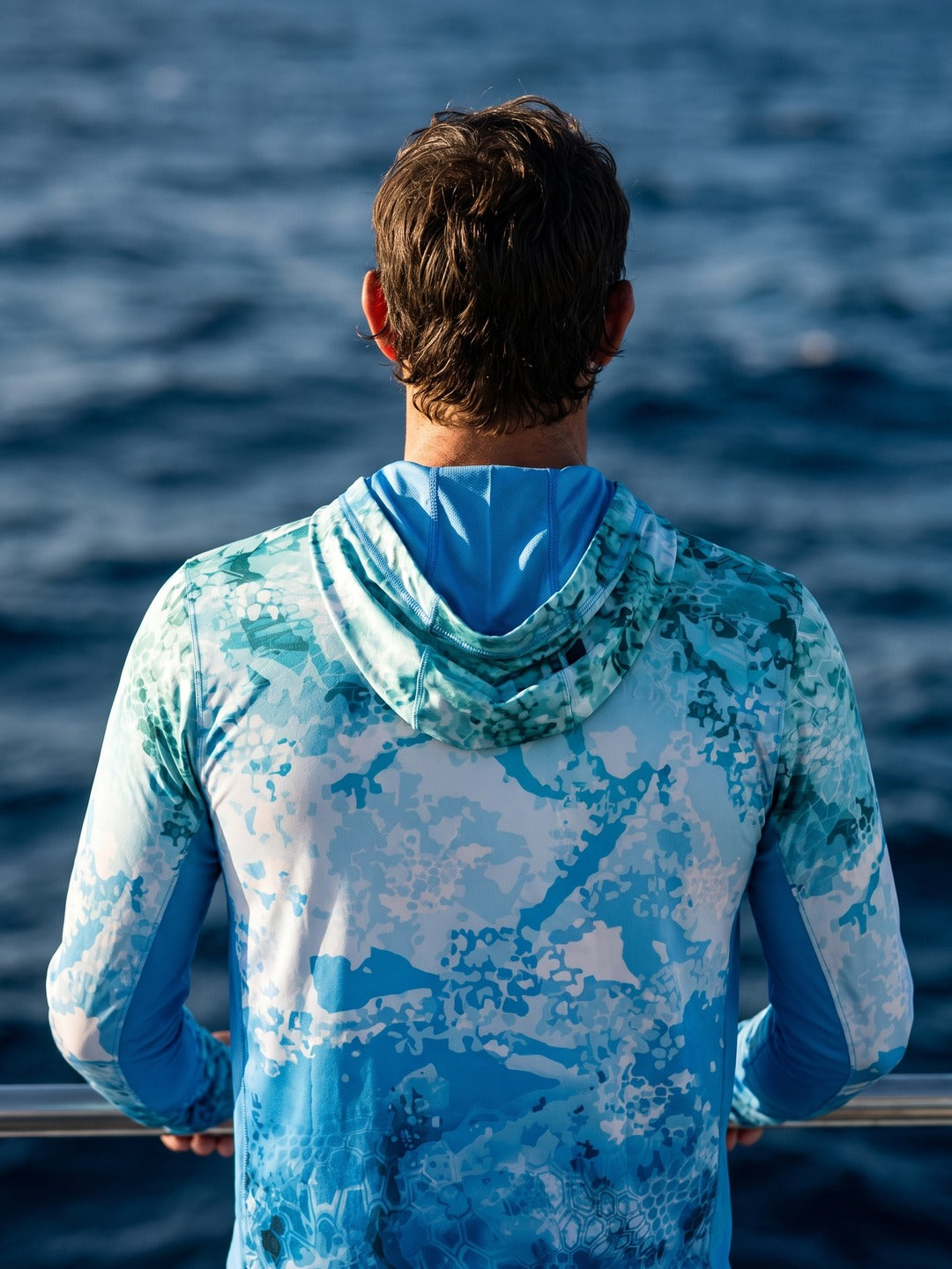 Back of the Ocean Pro Tech Hoodie worn on a boat, teal water-camo print with open ocean behind