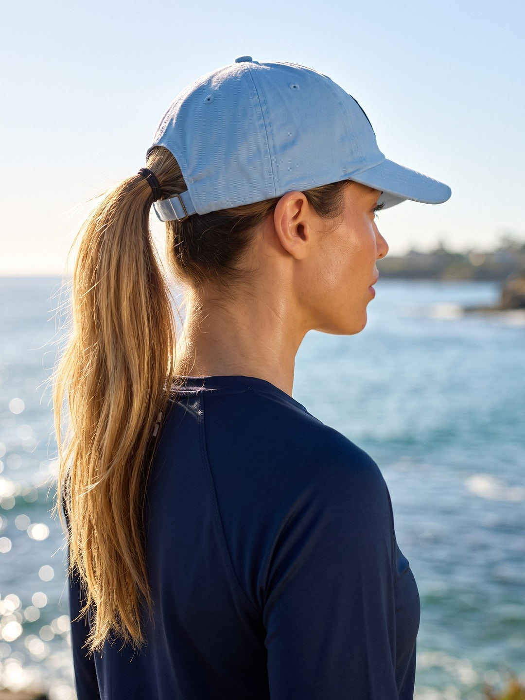 Woman with a ponytail wearing the light blue Round Logo Patch cap by the water